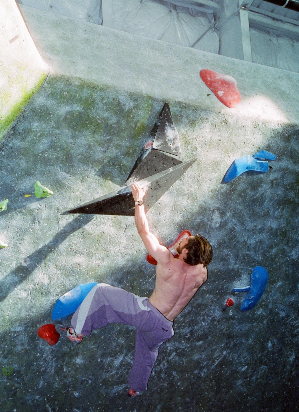 A man climbs a bouldering wall indoors, holding onto a black triangular climbing hold.