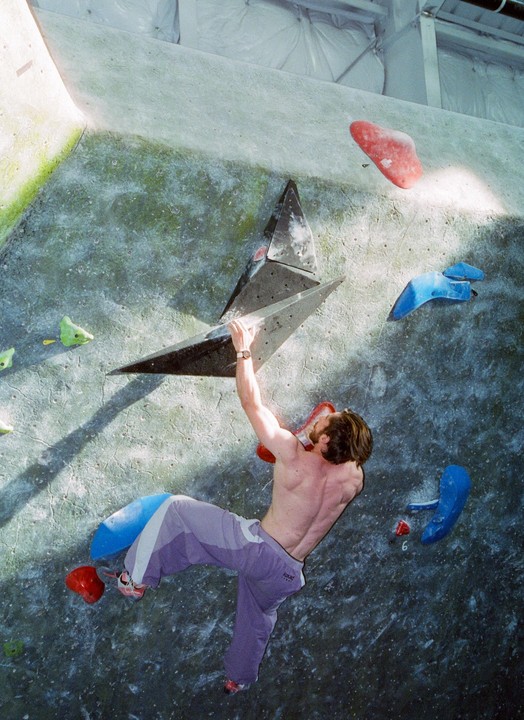 A man climbs a bouldering wall indoors, holding onto a black triangular climbing hold.