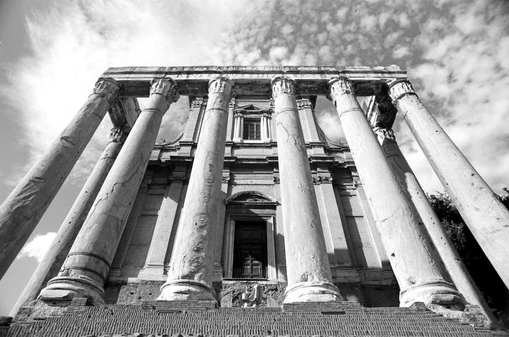 A low-angle view shows the ancient Temple of Antoninus and Faustina with its tall columns and a cloudy sky above.