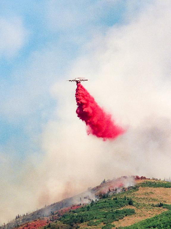 An airtanker drops red fire retardant over a smoky, forested hillside.