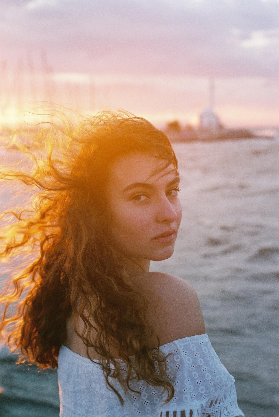 A woman with curly hair looks over her shoulder at the viewer, with the ocean and a distant building visible behind her.