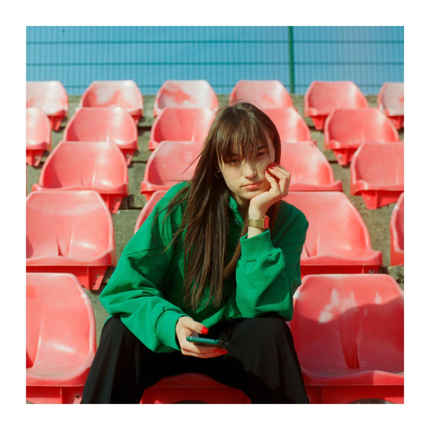 A woman in a green sweatshirt sits on a red stadium seat, holding a phone in her lap.