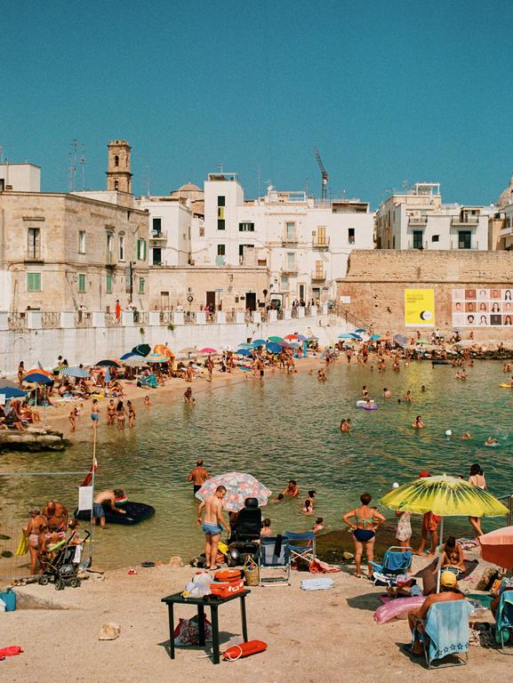 A crowded beach with people swimming and sunbathing is overlooked by white buildings and a bell tower under a clear blue sky.