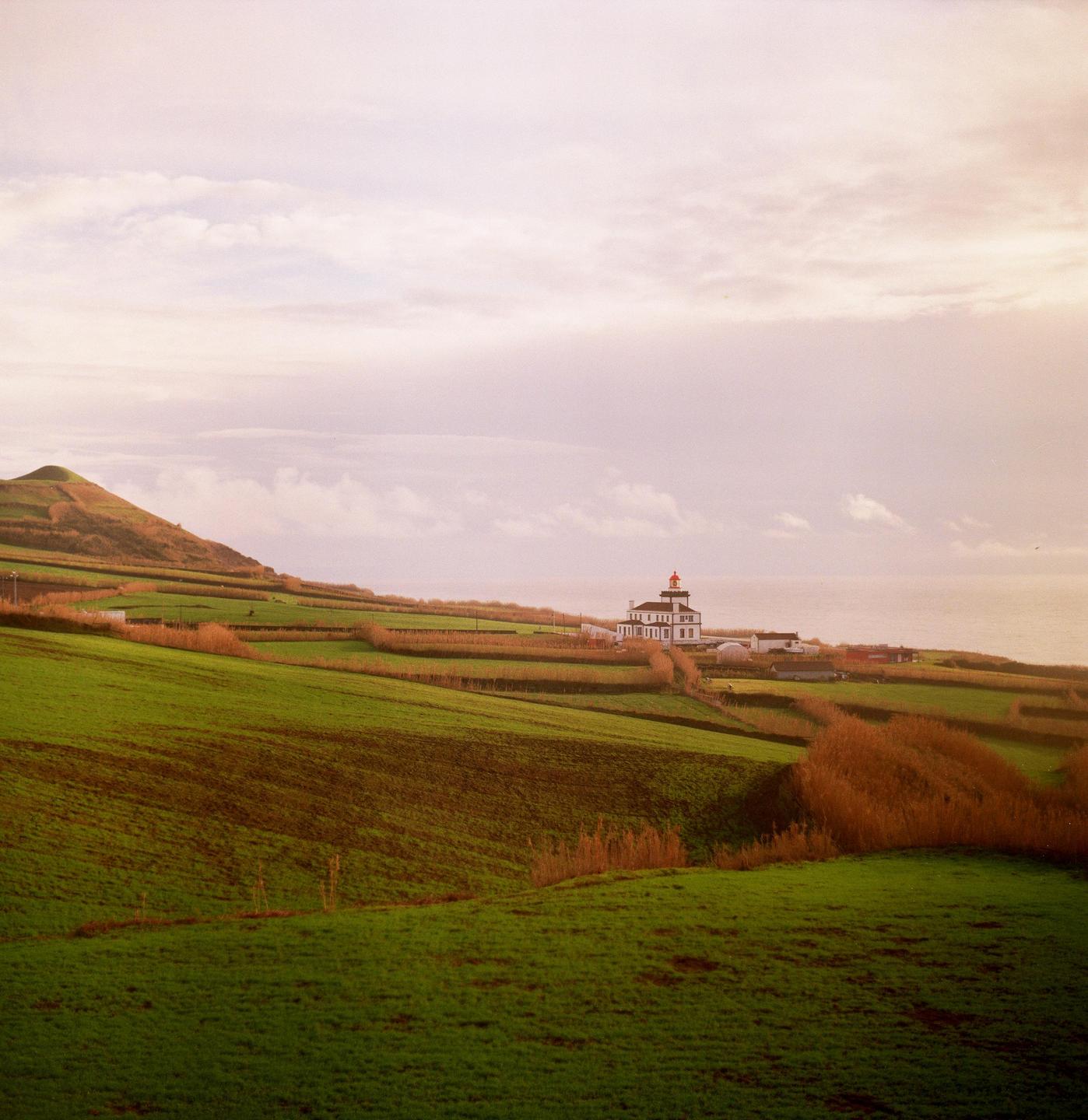 A white lighthouse with a red top stands on a green hillside overlooking the ocean under a cloudy sky.