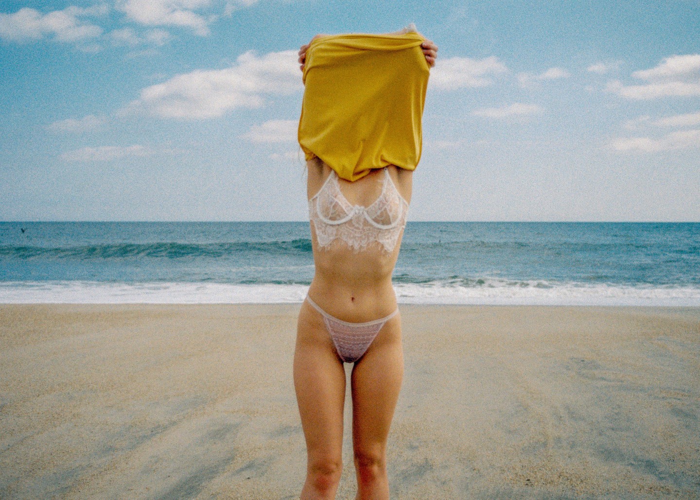 A woman in lingerie is pulling a yellow shirt over her head on a beach with the ocean in the background.