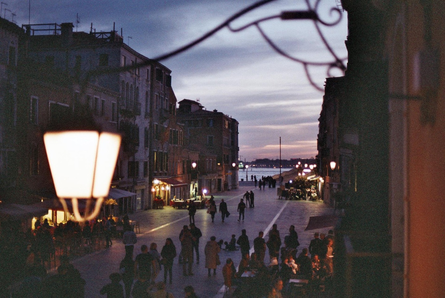 A street lamp illuminates a busy street with people and buildings at dusk, with a body of water visible in the distance.
