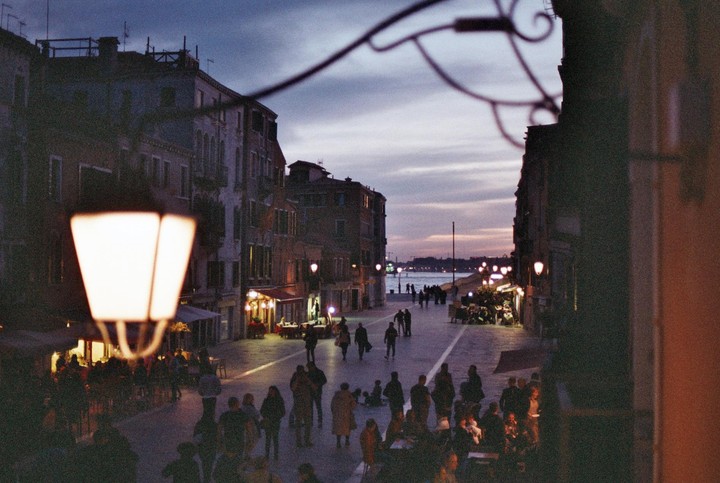 A street lamp illuminates a busy street with people and buildings at dusk, with a body of water visible in the distance.