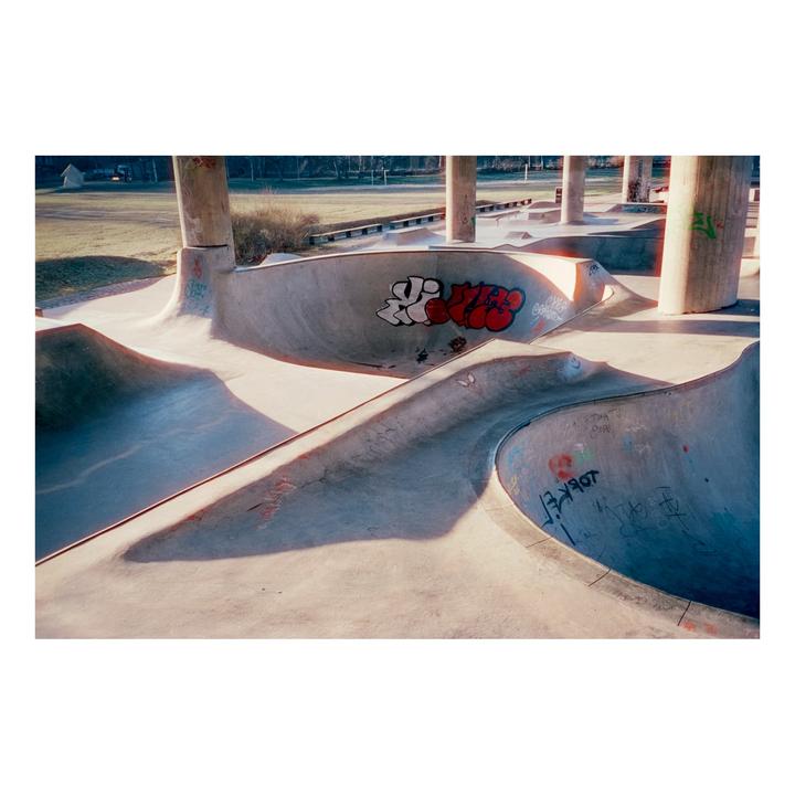 A skatepark with concrete bowls and pillars is illuminated by sunlight, casting long shadows.