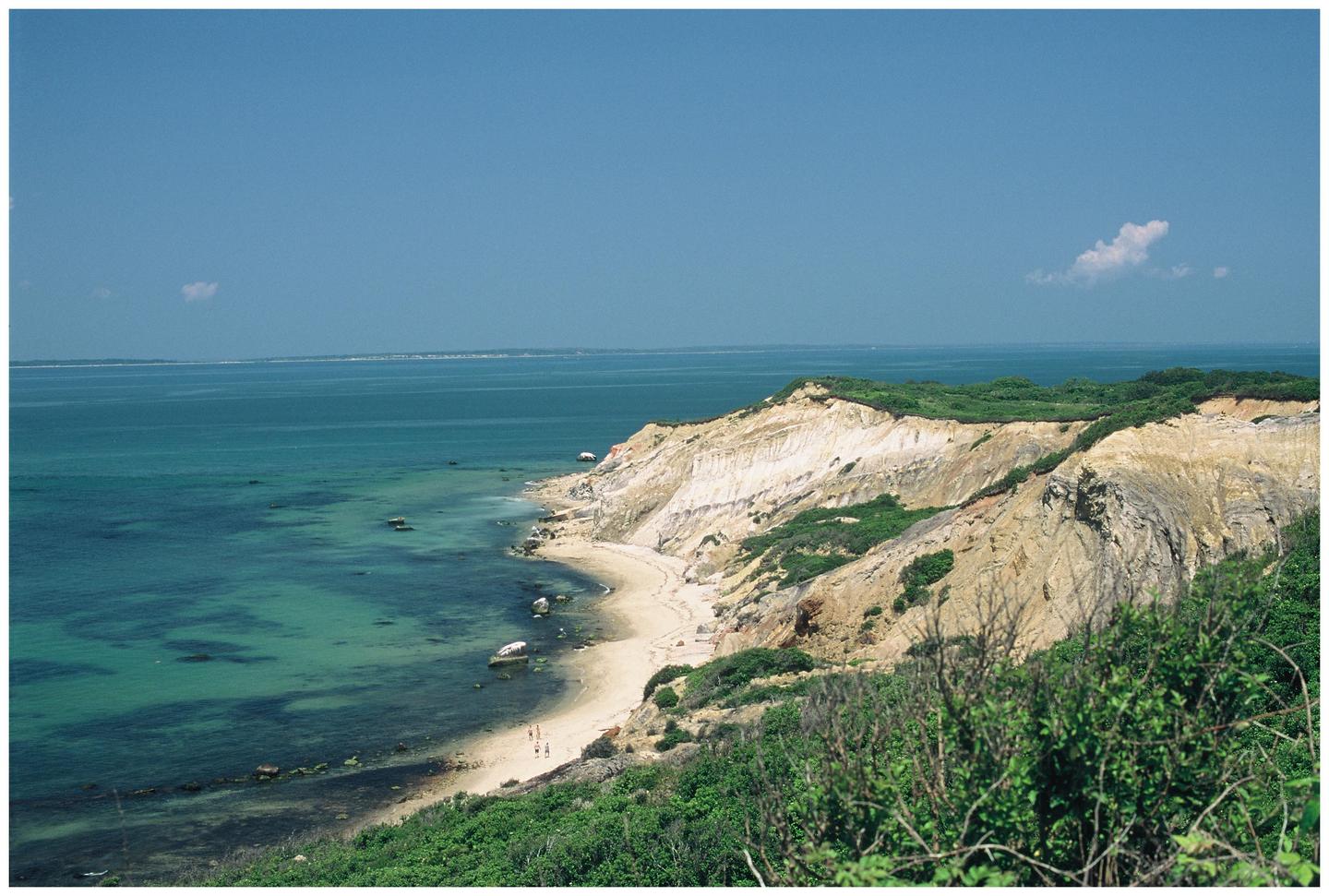 A scenic view shows a sandy beach nestled at the base of a cliff, with clear blue ocean water stretching to the horizon under a bright sky.