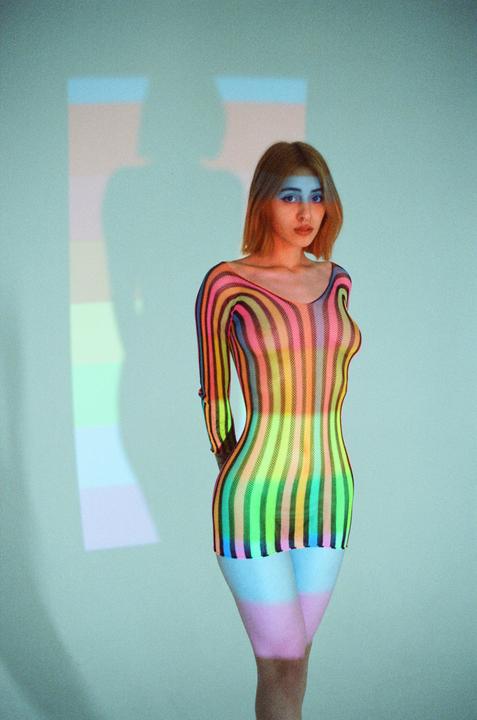 A woman in a colorful striped dress stands in front of a light blue wall with a colorful shadow.