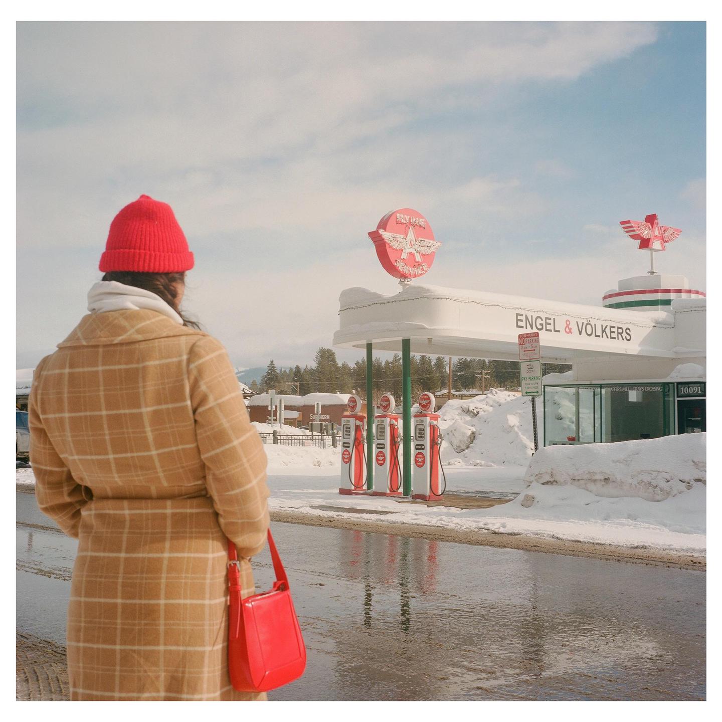 A woman in a plaid coat and red hat stands on a wet street looking at a gas station covered in snow.