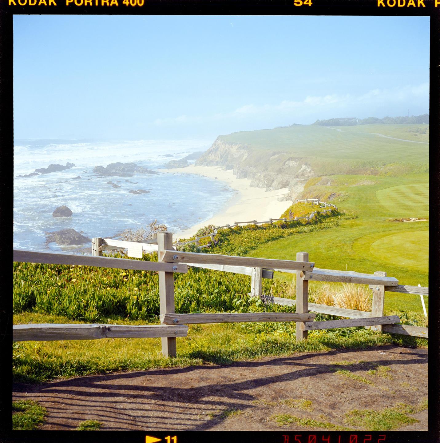 A wooden fence overlooks a sandy beach and a golf course under a hazy sky.