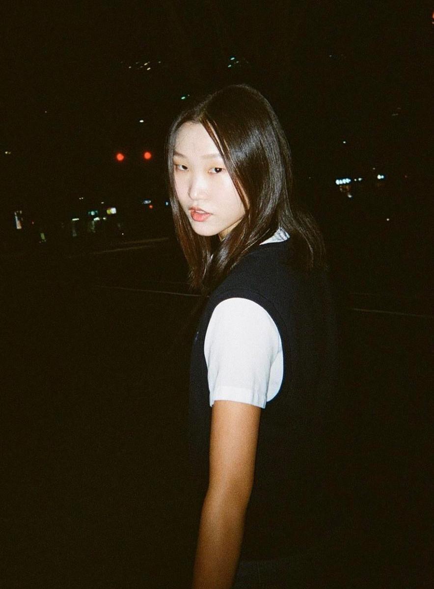 A young woman with long hair looks over her shoulder at night with city lights in the background.