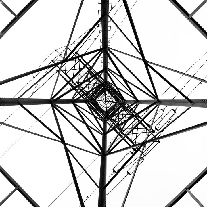 A low-angle view of a power line tower shows its symmetrical structure against a white sky.