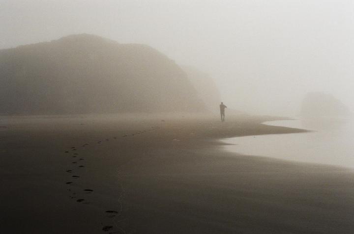 A man walks along a foggy beach with mountains in the background.