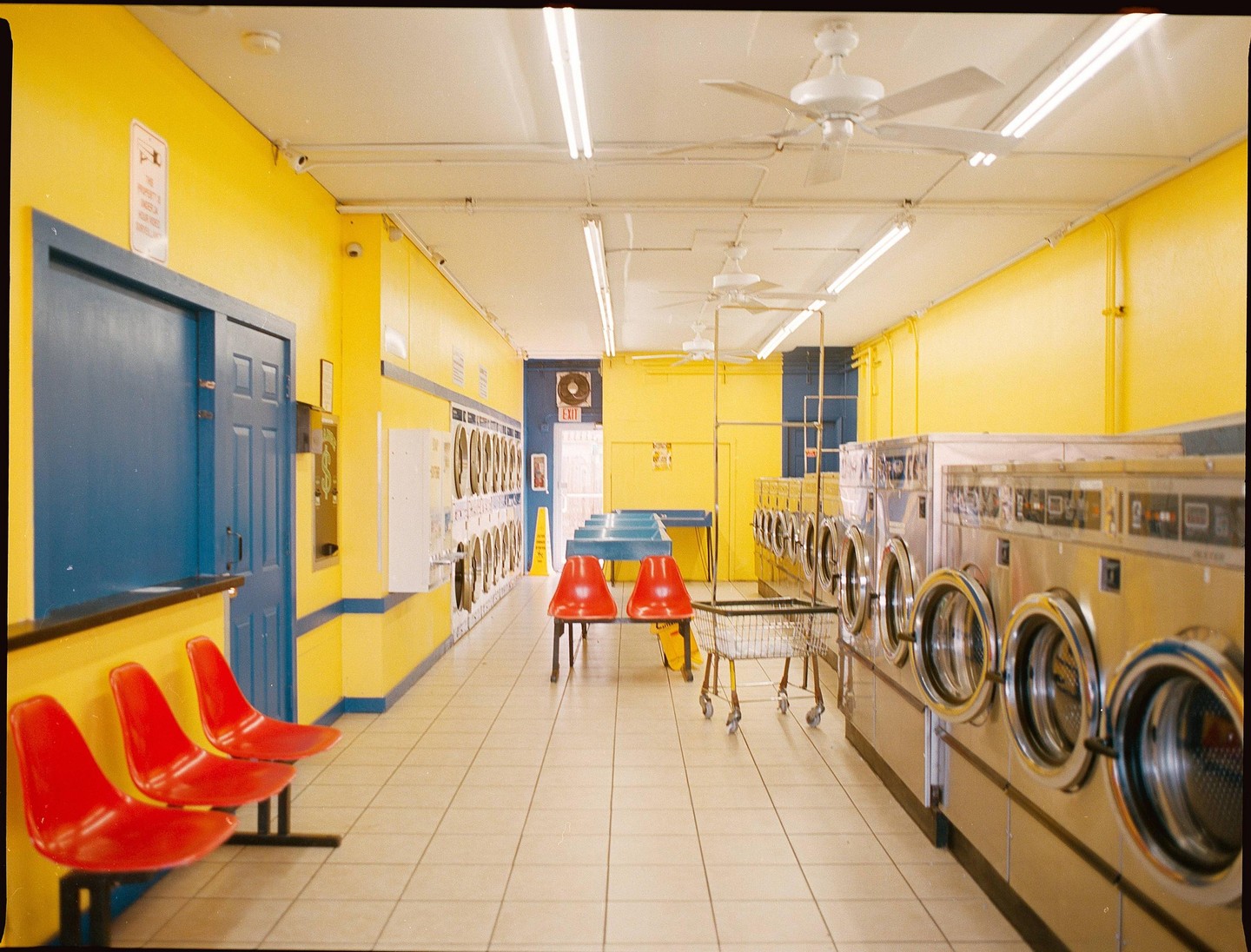 A brightly lit laundromat with yellow walls and blue trim features rows of washing machines, red chairs, and ceiling fans.