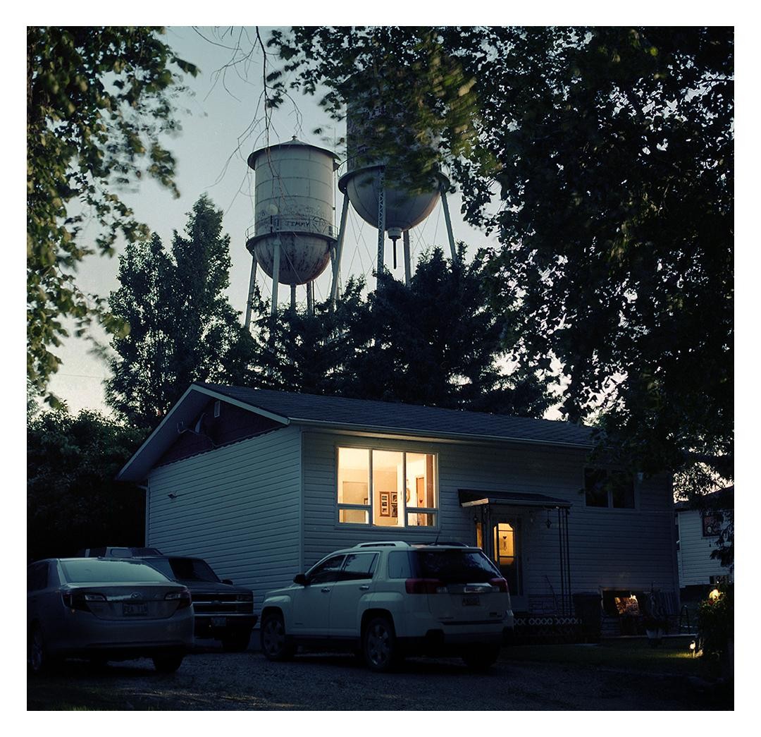 A house with illuminated windows and parked cars is visible beneath two water towers and trees at dusk.