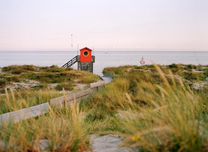 A red lifeguard hut stands on a sandy dune overlooking the sea, with a wooden walkway leading towards it.