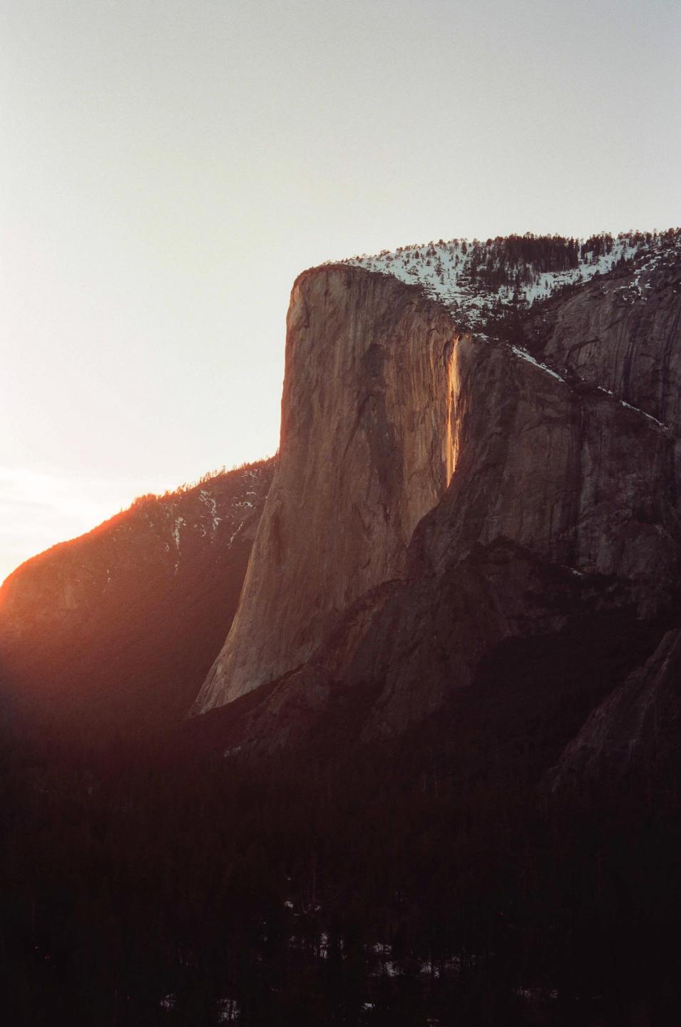 A large rock formation is illuminated by a golden light, with snow-capped trees visible on its peak and a dark forest below.