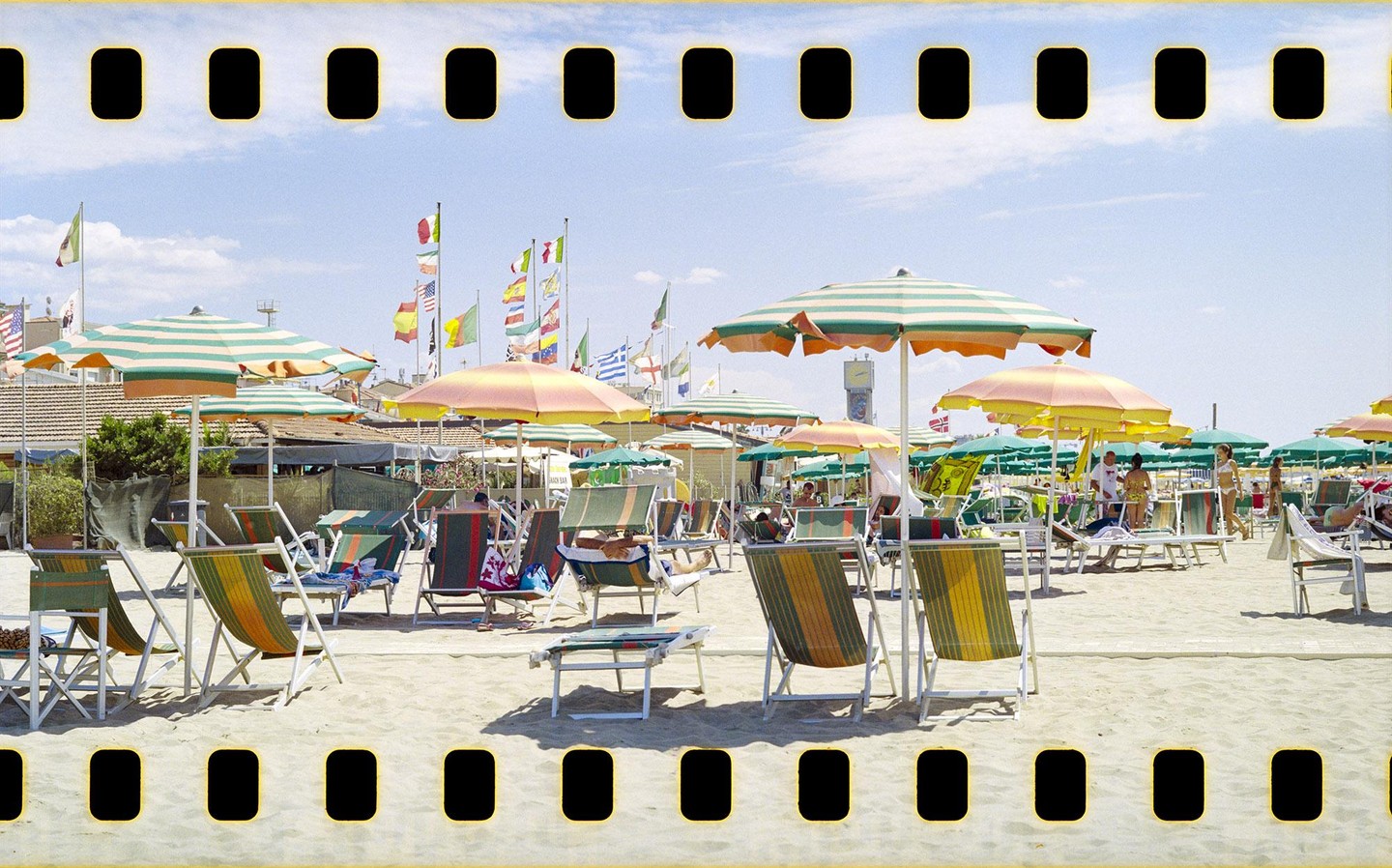 A sandy beach is filled with striped umbrellas and lounge chairs under a bright sky with many flags flying in the background.