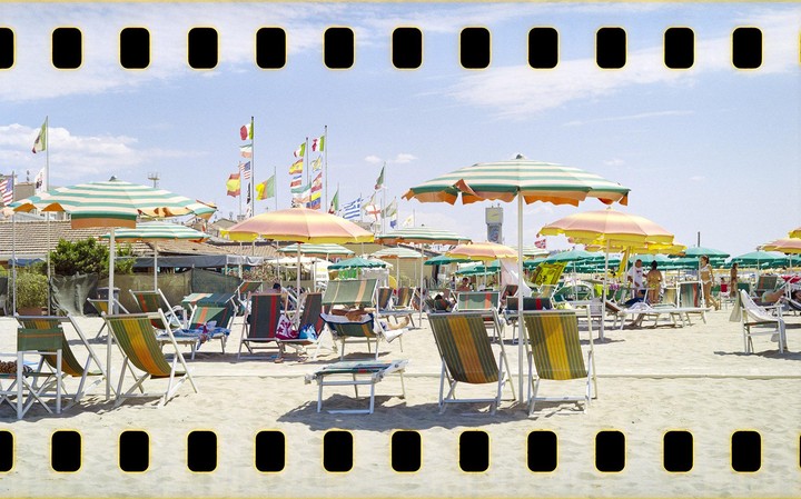 A sandy beach is filled with striped umbrellas and lounge chairs under a bright sky with many flags flying in the background.