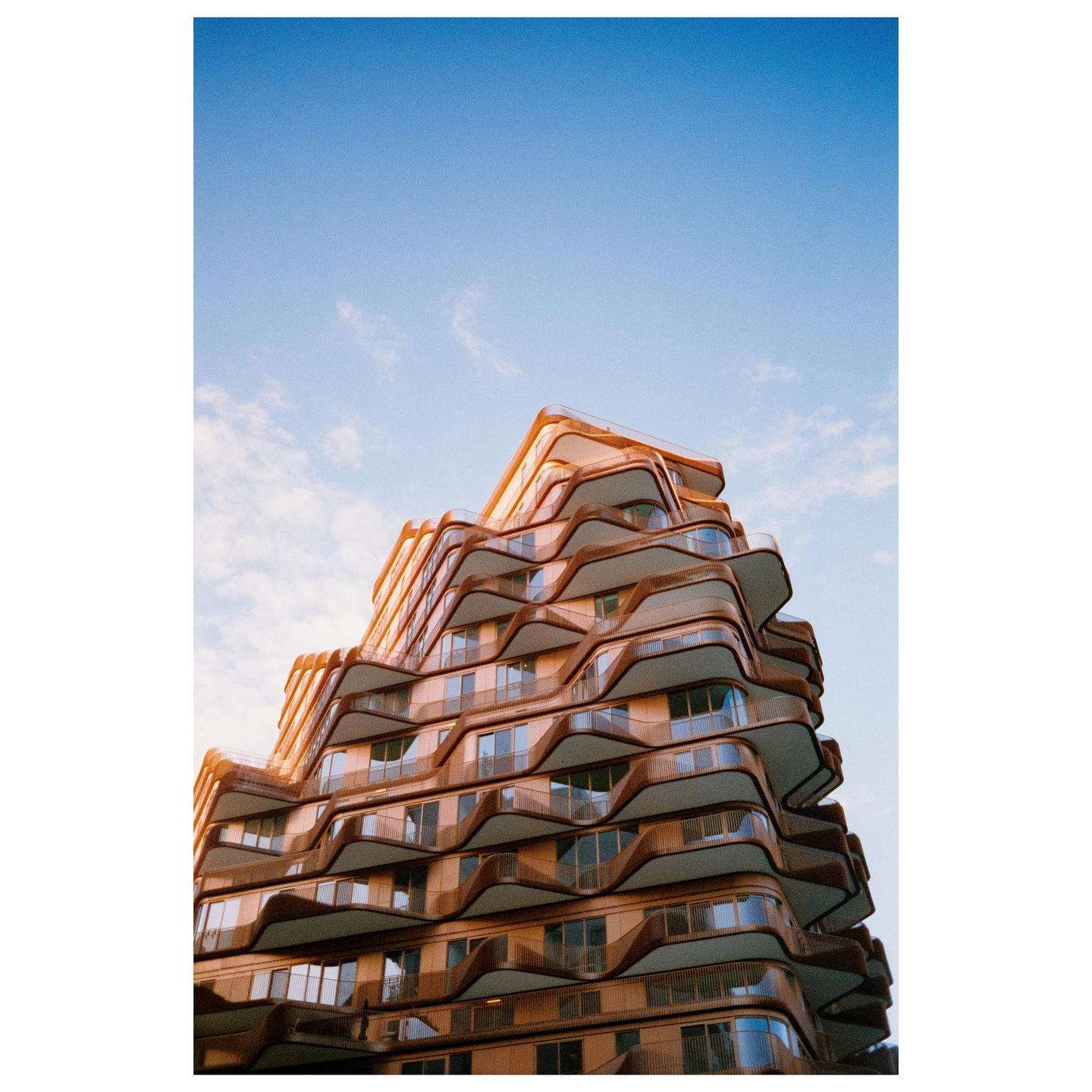 A modern building with wavy balconies and a copper facade stands against a clear blue sky.