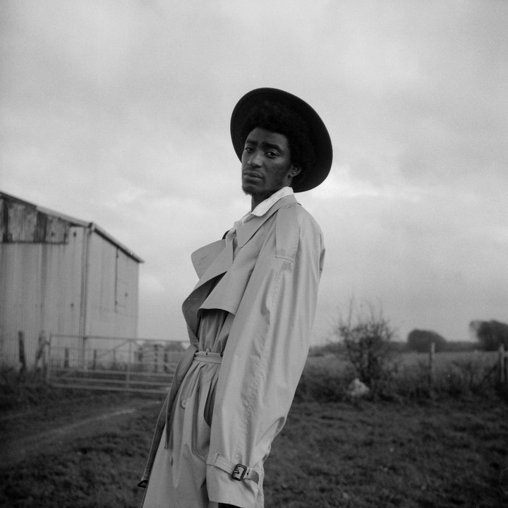 A man in a trench coat and hat stands in a field with a building in the background.
