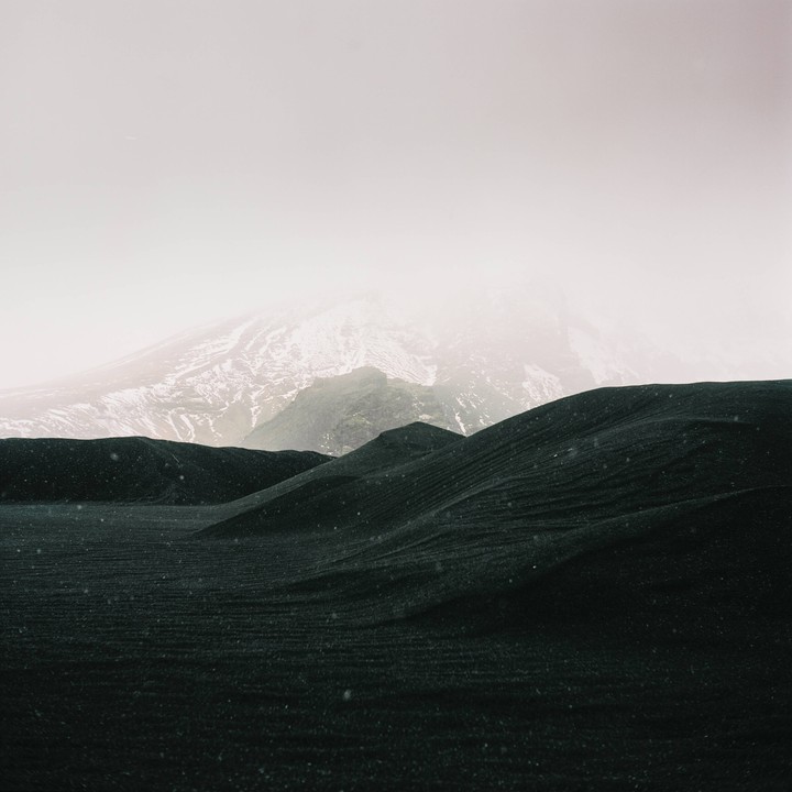 Snow falls on dark, rolling hills with a snow-capped mountain in the background.