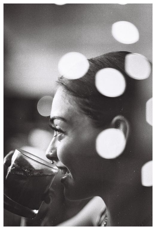 A woman with a nose piercing drinks from a glass, with bokeh circles superimposed over her head.