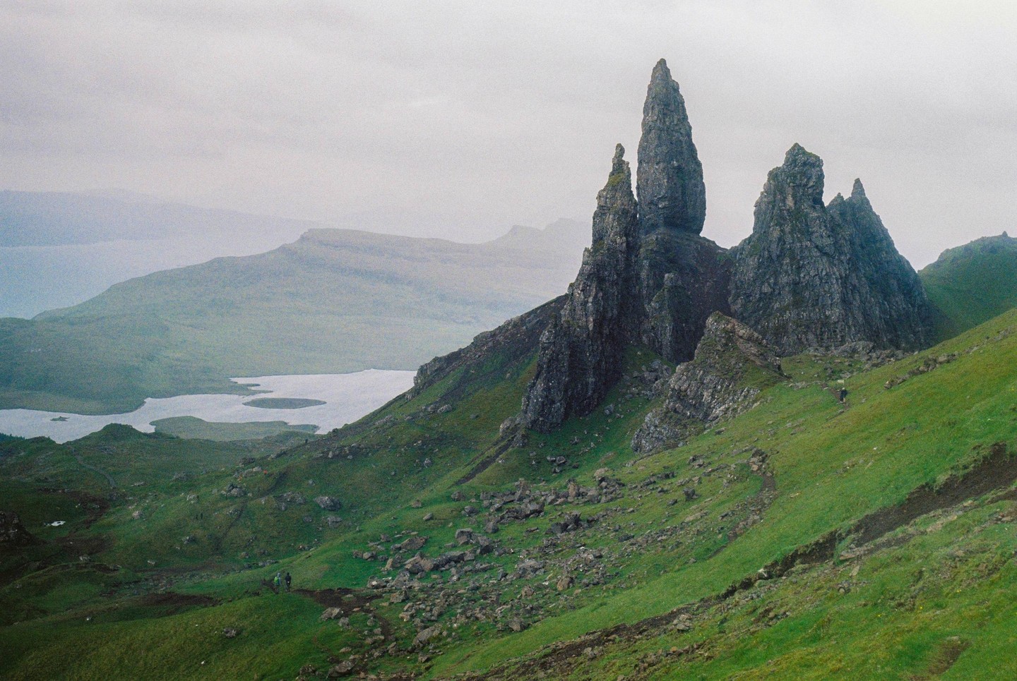 A person walks on a path through a green, rocky landscape with a large rock formation and a lake in the distance.