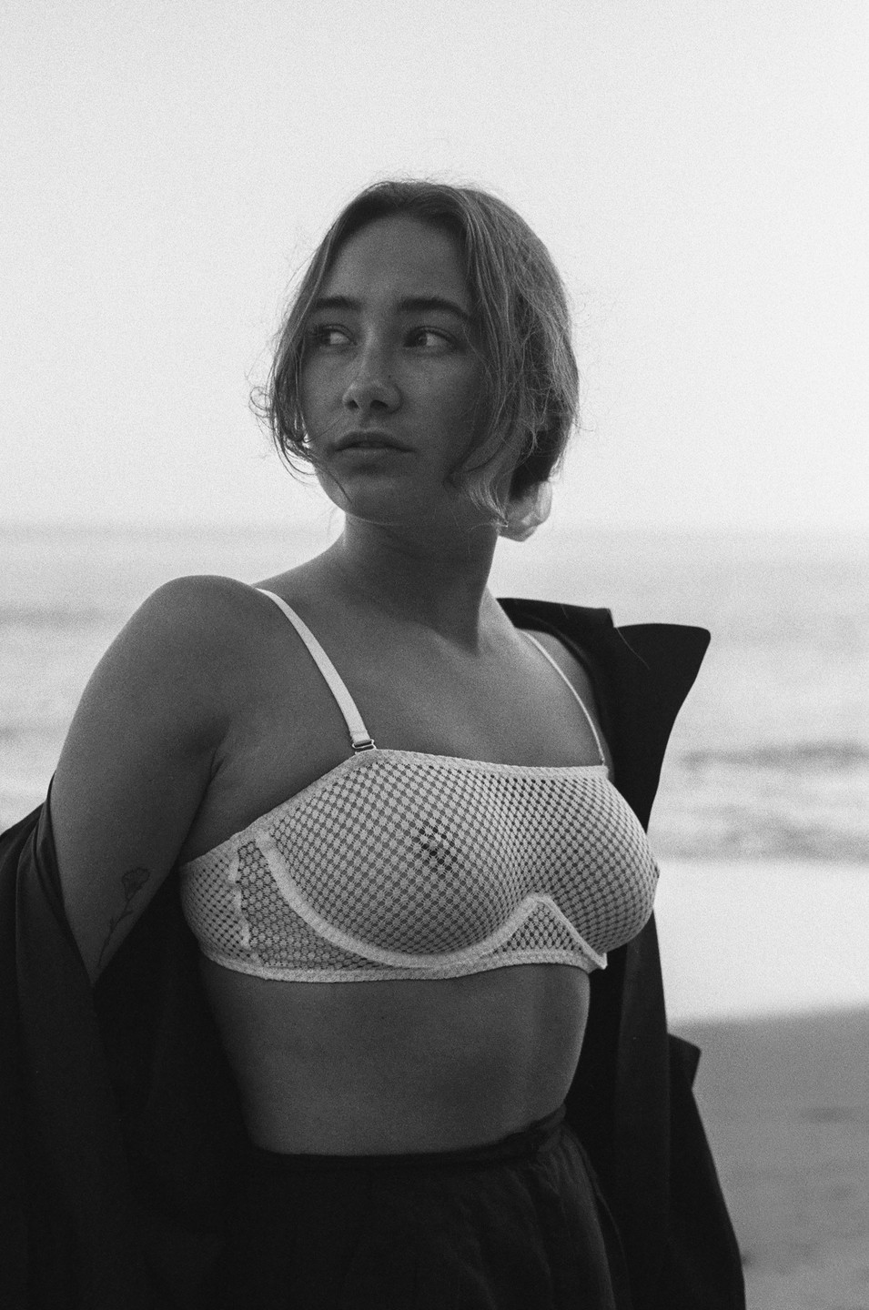 A woman in a mesh bra and dark jacket stands on a beach looking away from the camera.