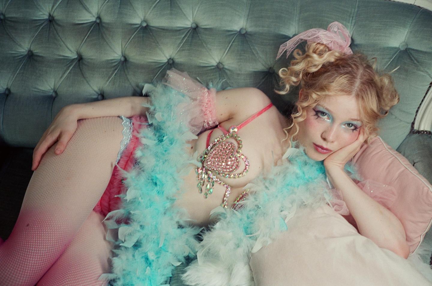 A woman with curly blonde hair and elaborate makeup reclines on a tufted sofa, adorned with a feather boa and a sparkling heart-shaped pendant.