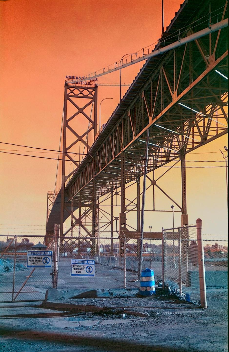 A large metal bridge with a sign that reads "Ambassador Bridge" is visible against an orange sky, with a chain-link fence and two "restricted area" signs in the foreground.