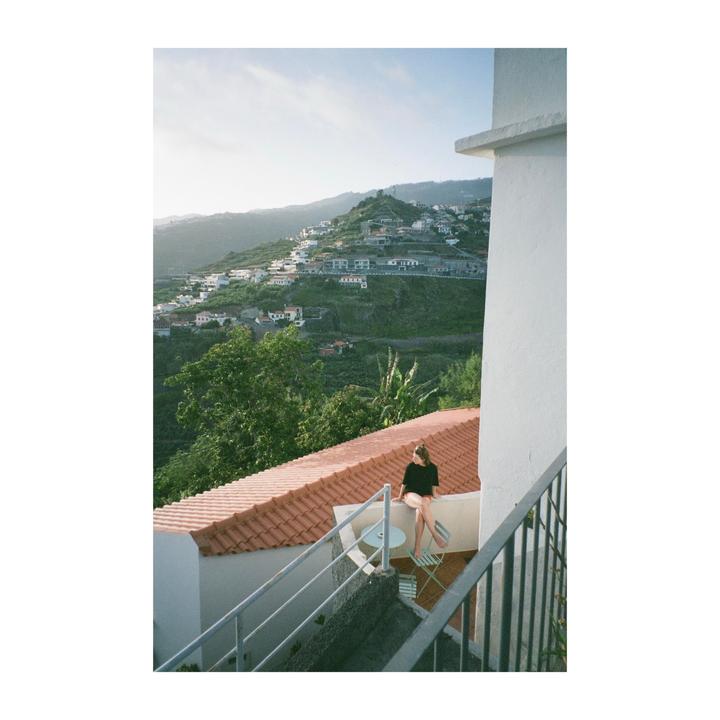 A woman sits on a balcony overlooking a village built into a hillside.