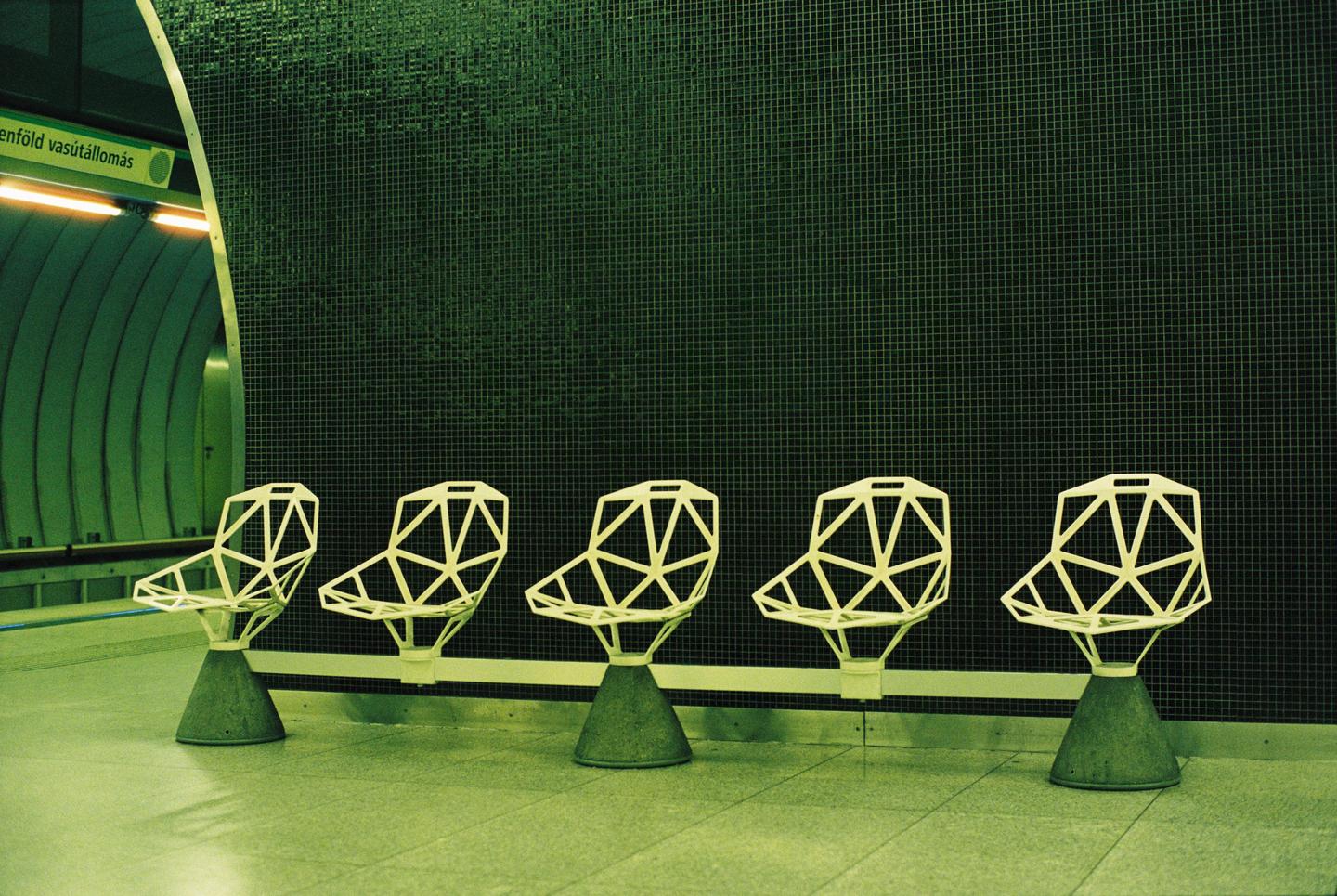 Five geometric chairs are lined up against a dark tiled wall in a subway station.