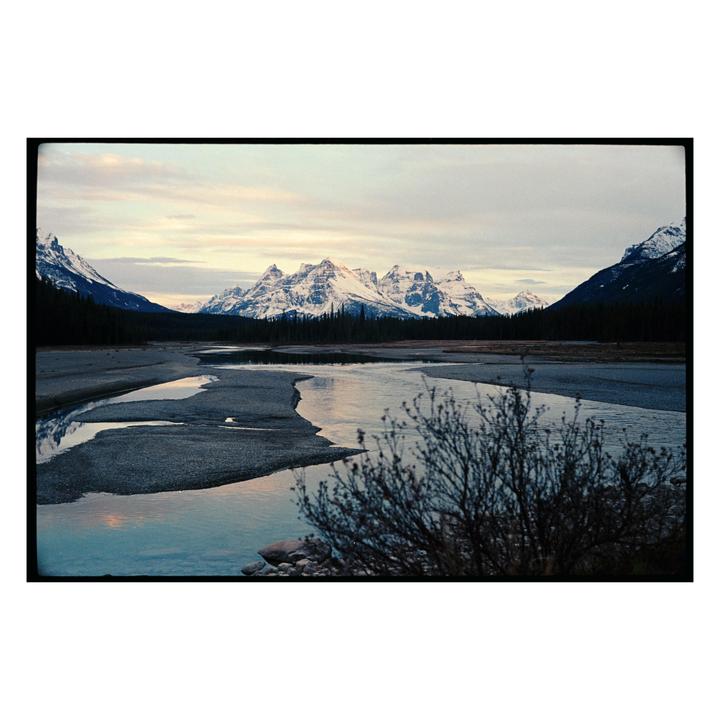 A river flows through a valley with snow-capped mountains and evergreen trees under a cloudy sky.