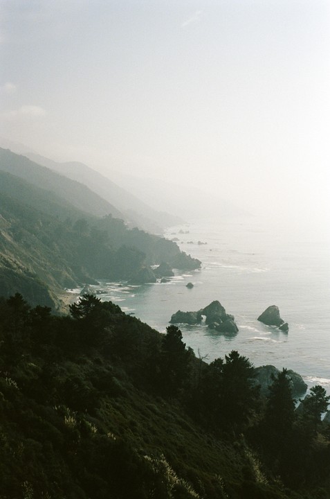 A hazy view of the California coast shows a rugged coastline with mountains, trees, and rock formations in the ocean.