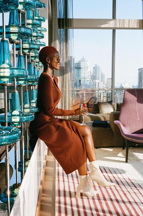 A woman in a brown dress sits on a ledge next to a large blue glass sculpture, holding a drink and looking out a window at a city skyline.