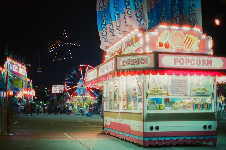 A brightly lit popcorn and drinks stand is visible at a fair at night, with a Ferris wheel and other attractions in the background.