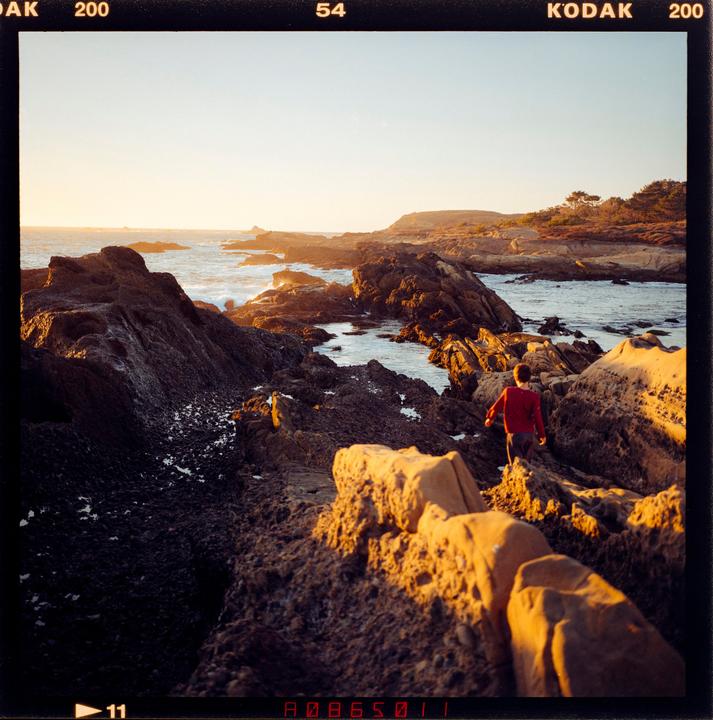 A boy walks along a rocky coastline at sunset.