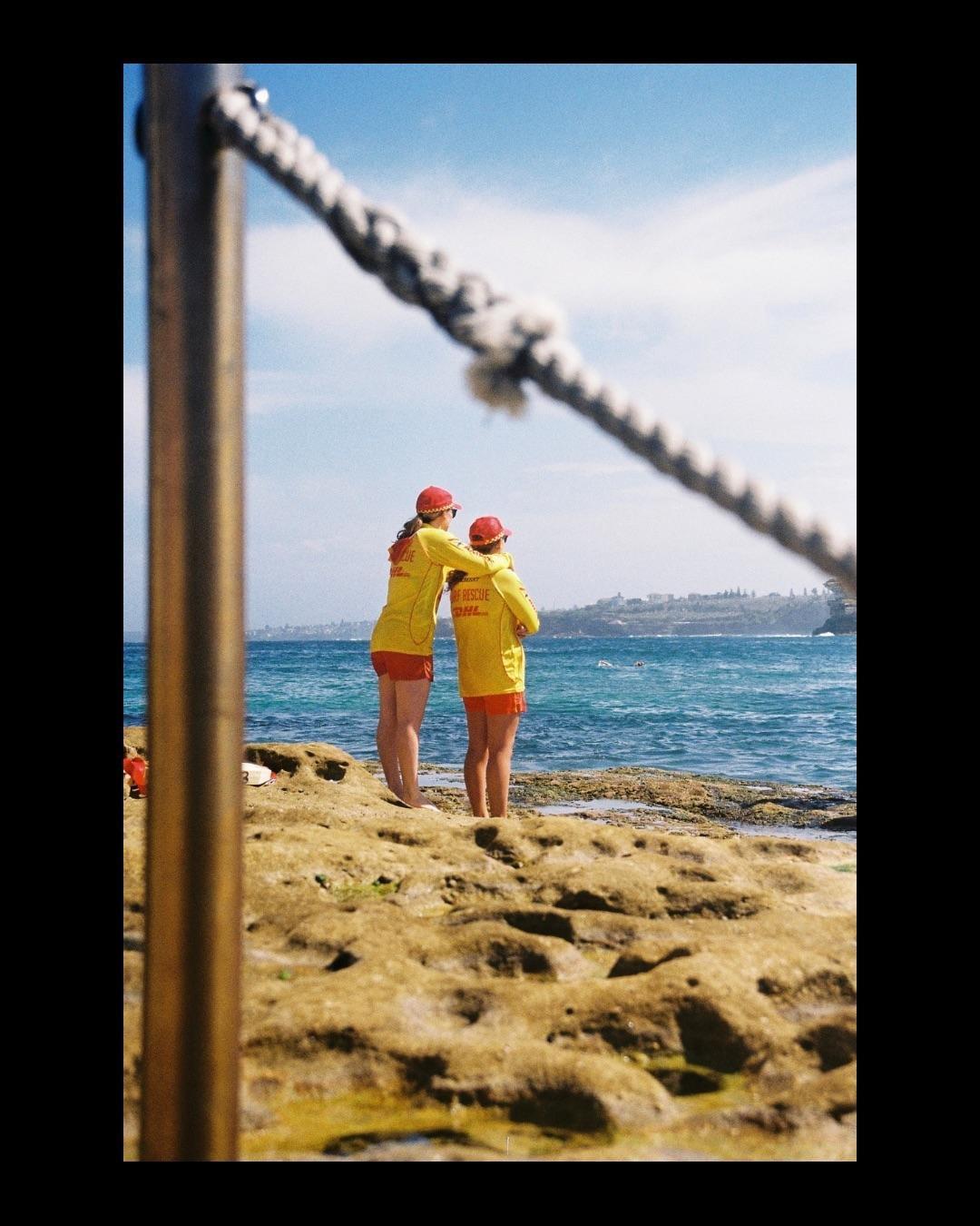 Two women in yellow shirts and red hats stand on a rocky shore looking out at the ocean.