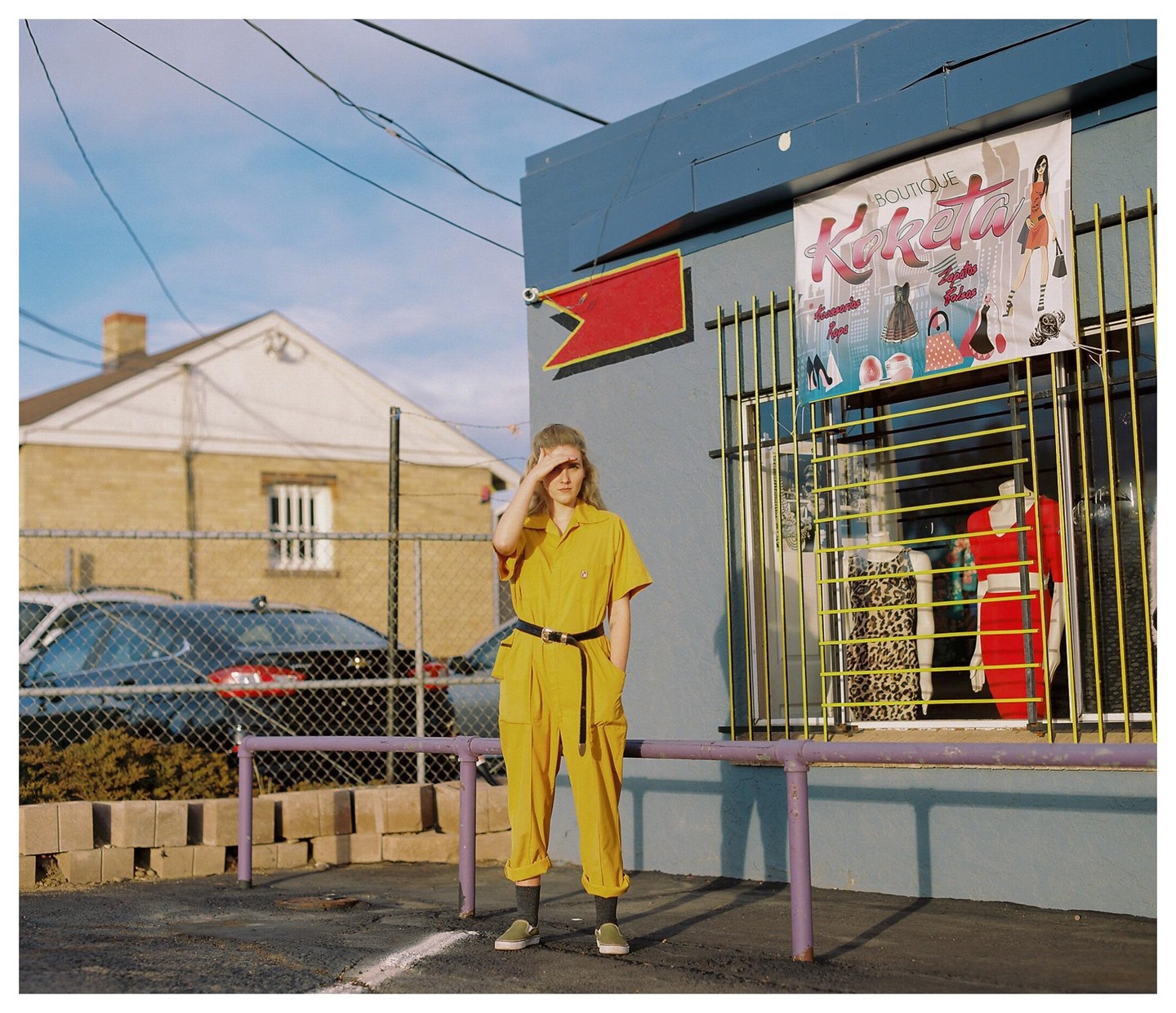 A woman in a yellow jumpsuit shields her eyes from the sun while standing in front of a boutique.