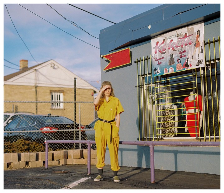A woman in a yellow jumpsuit shields her eyes from the sun while standing in front of a boutique.
