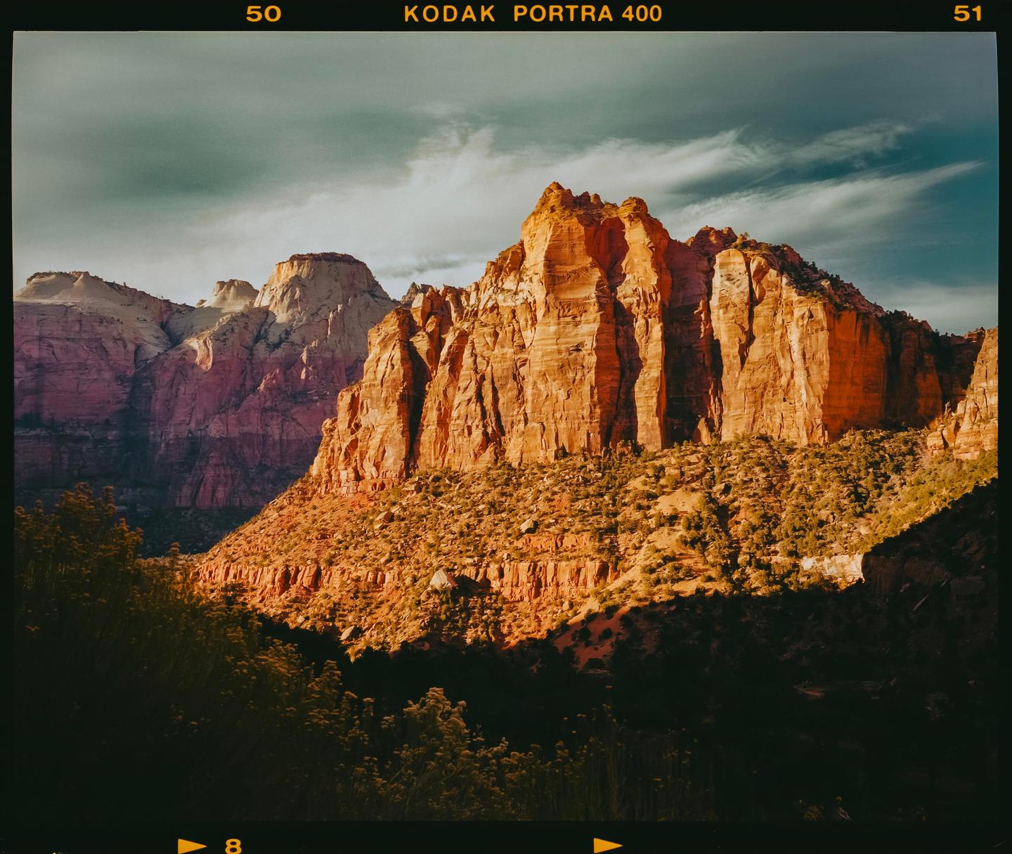 A large mountain range with red rock formations is illuminated by sunlight under a cloudy sky.