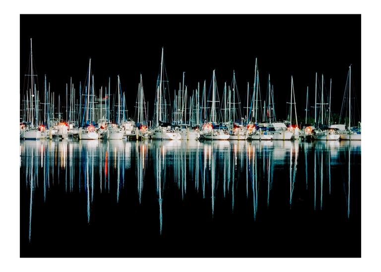 Many sailboats are docked at night, their masts and lights reflecting in the water.
