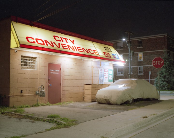 A covered car is parked next to a convenience store with a bright yellow awning at night.