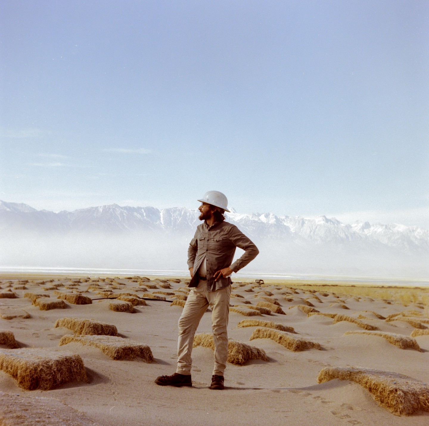 A man wearing a hard hat stands in a desert landscape with mountains in the background.