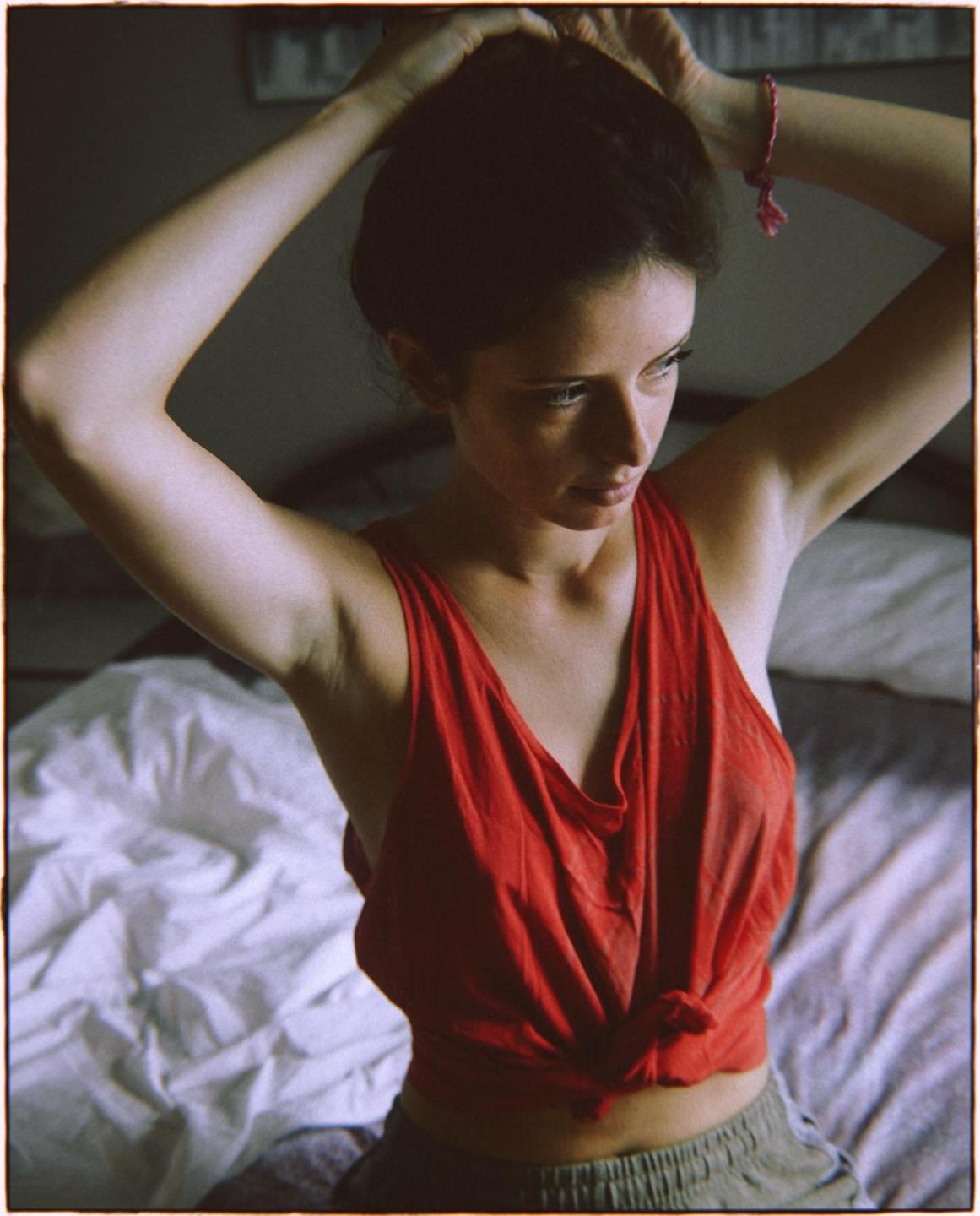 A woman in a red top is tying her hair up while sitting on a bed.