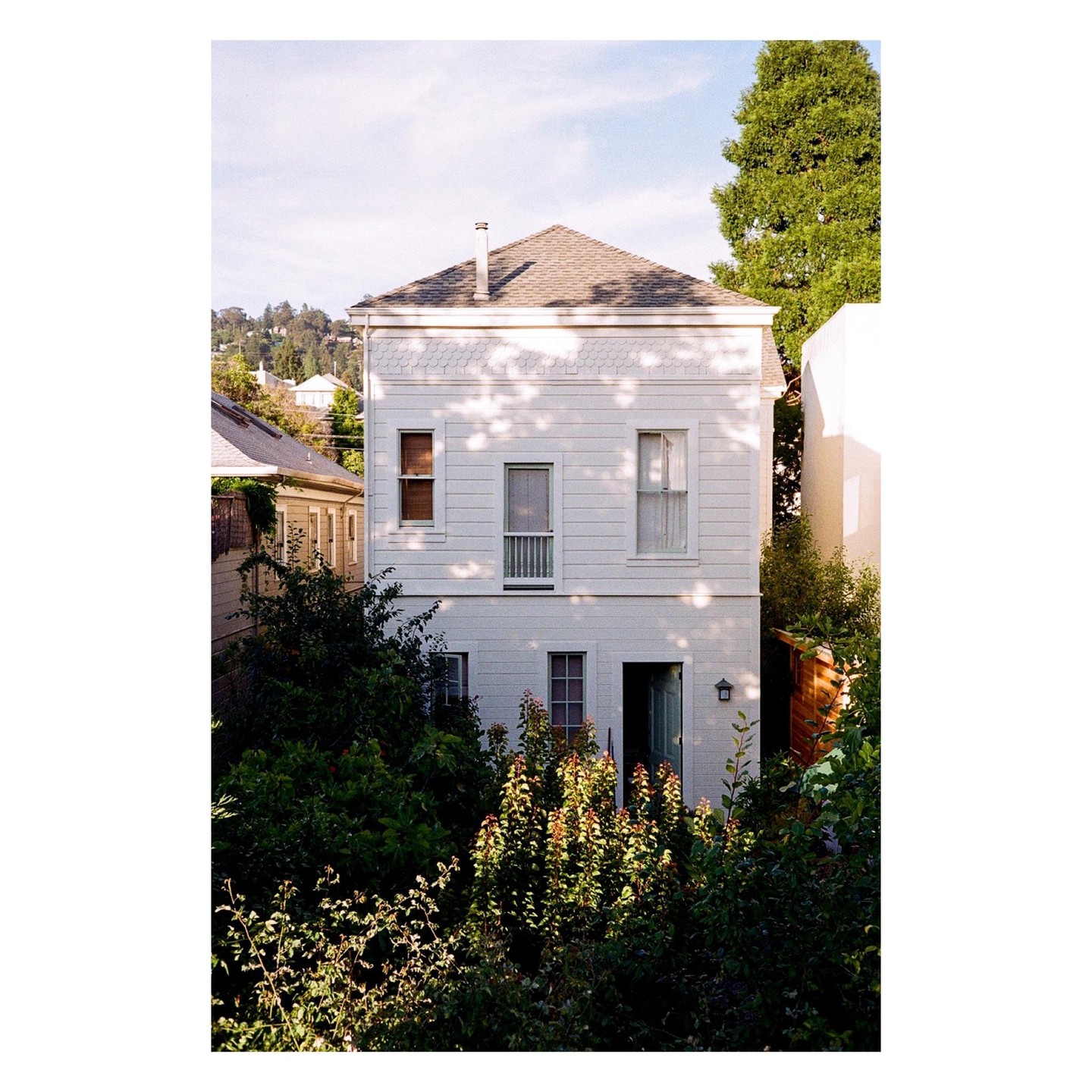 A white house with a gray roof is surrounded by green foliage under a bright sky.