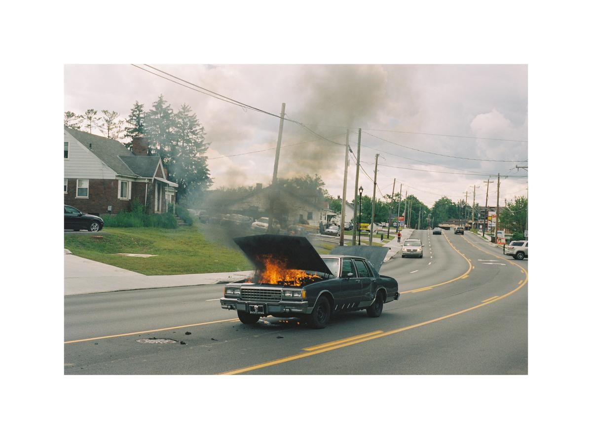 A car with its hood open is on fire on the side of a road with houses and trees in the background.